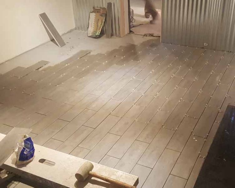 A man installing long, gray tiles on a floor in a spacious room, with construction tools scattered around.