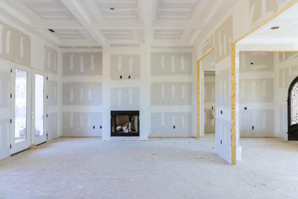 A large room under construction with unfinished drywall and exposed beams, large windows, and a fireplace structure, illustrating the early stages of a home build or renovation project.